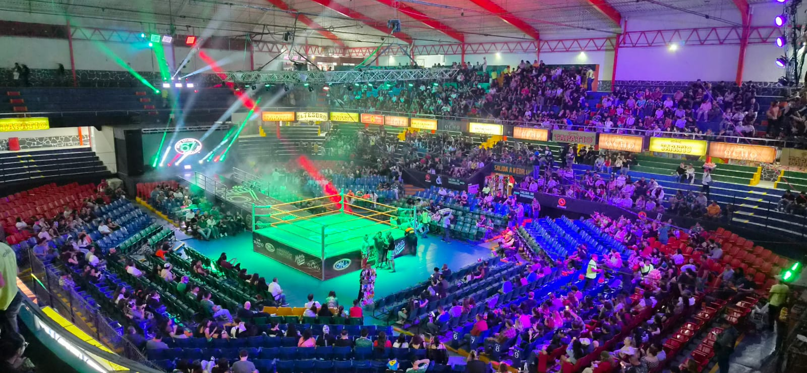 A lucha libre arena filled with spectators, with a brightly lit wrestling ring at the center.