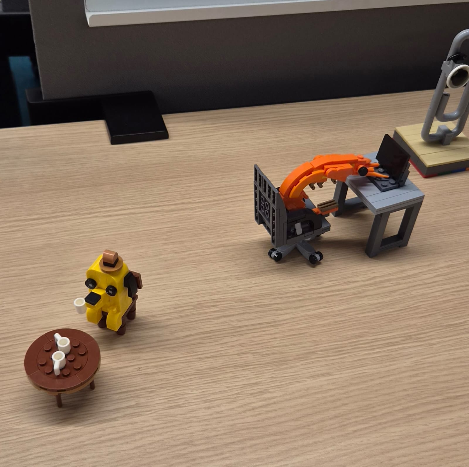 A LEGO dog with coffee beside a desk setup and a LEGO mechanical arm.